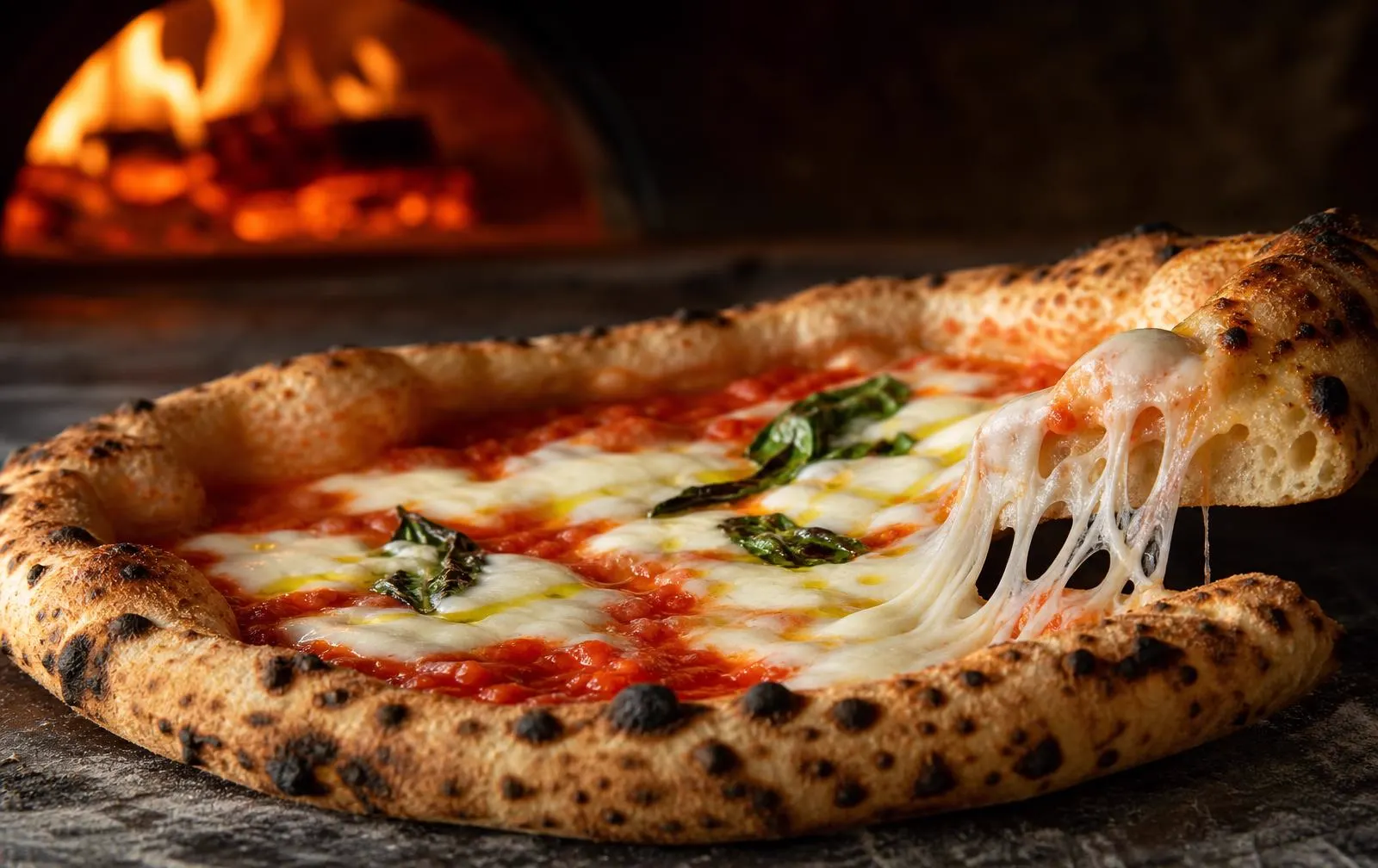 Pizza margherita appena sfornata con cornicione alveolato e mozzarella filante davanti al forno a legna